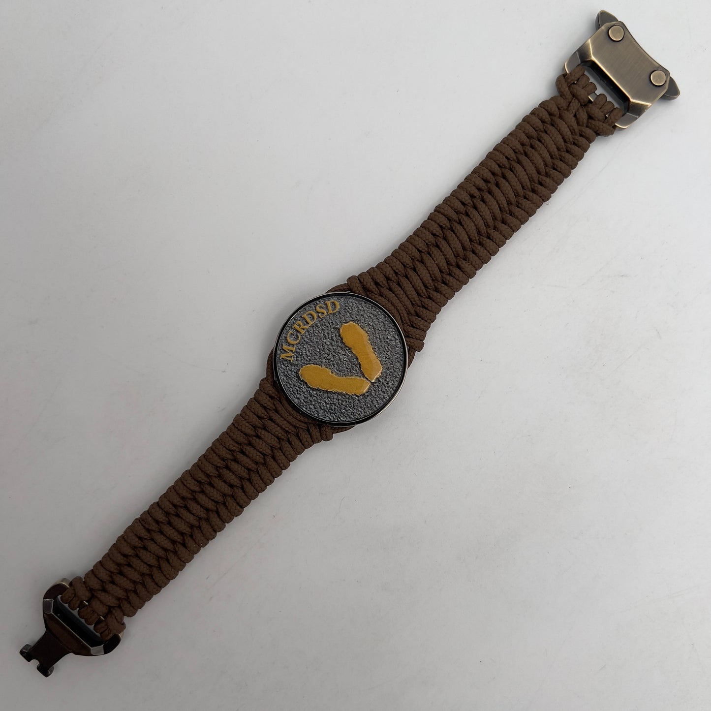 Thin, narrow, flexible and comfortable braided United States Marine Corps Recruit Depot Yellow Footprints bracelet featuring a black nickel plated UV enamel painted medallion replica of an actual set of USMC recruit depot footprints, a gold Cobra style tactical buckle and woven with Coyote Brown type-1 paracord.
