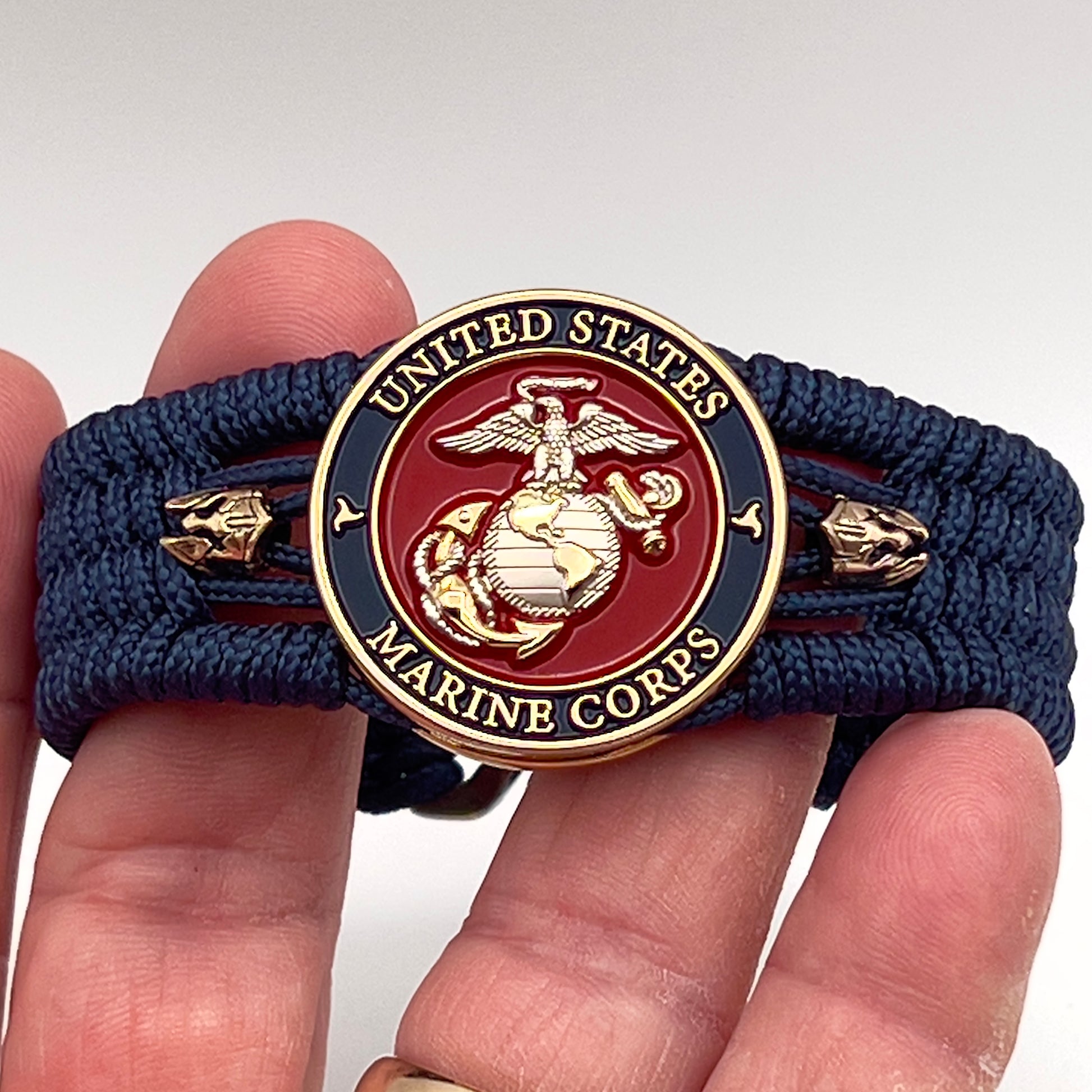 Thin, medium width, flexible and comfortable braided United States Marine Corps bracelet featuring a 24K gold and silver plated USMC officer Eagle Globe and Anchor medallion, two brass Spartan helmets, a gold Cobra style tactical buckle and woven with Navy Blue type-1 paracord.