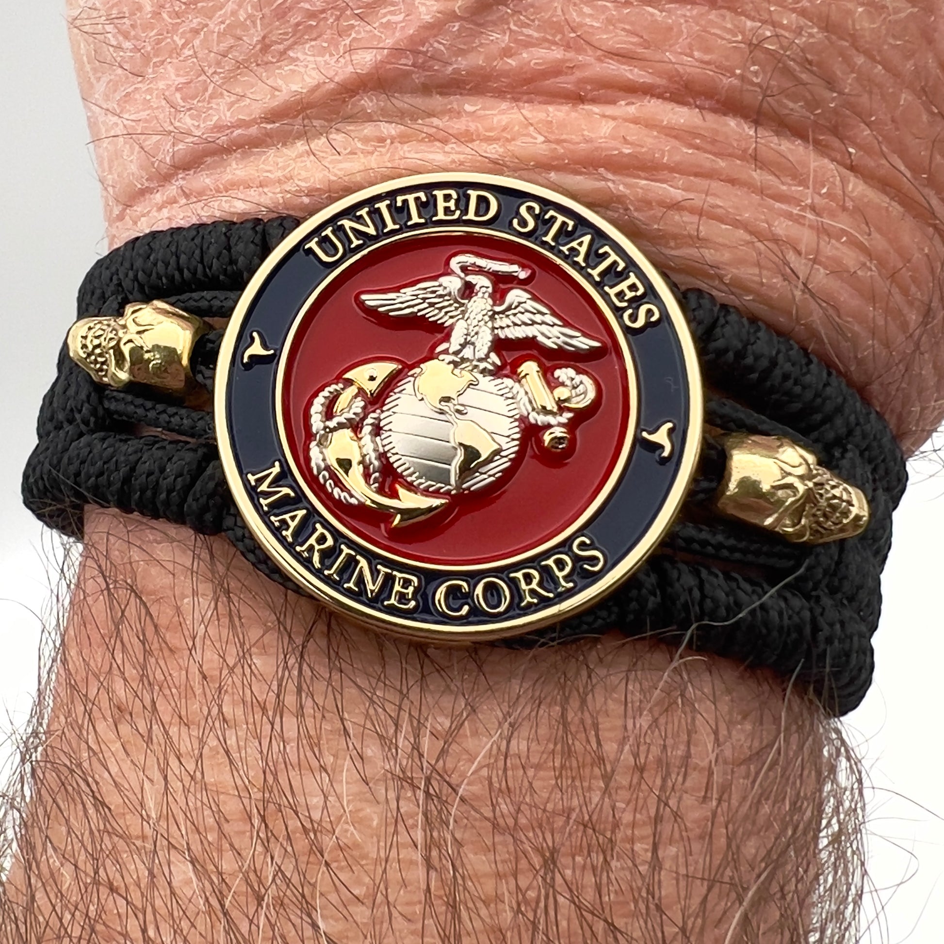 Thin, medium width, flexible and comfortable braided United States Marine Corps bracelet featuring a 24K gold and silver plated USMC officer Eagle Globe and Anchor medallion, two gold plated skulls, a black Cobra style tactical buckle and woven with black type-1 paracord.