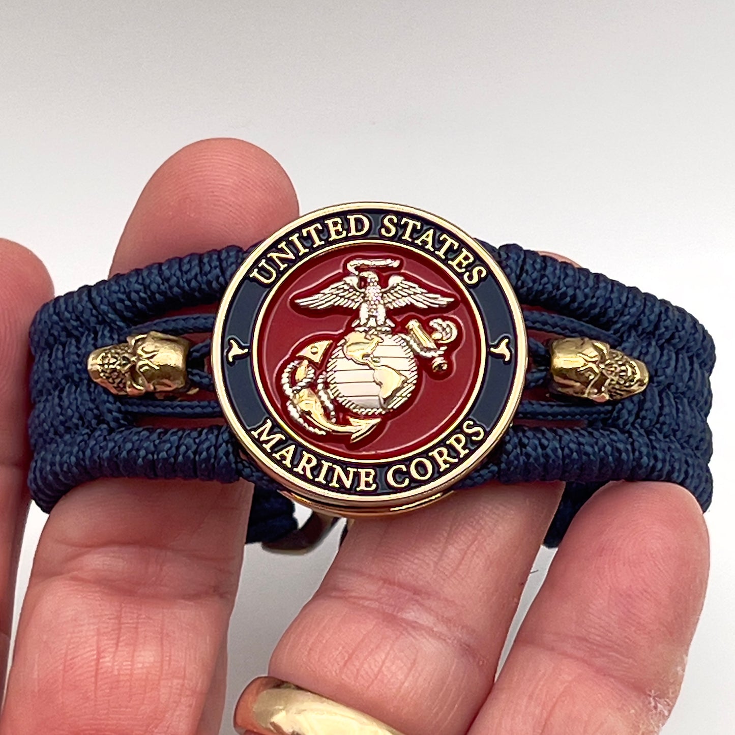 Thin, medium width, flexible and comfortable braided United States Marine Corps bracelet featuring a 24K gold and silver plated USMC officer Eagle Globe and Anchor medallion, two gold micro skulls, a gold Cobra style tactical buckle and woven with Navy blue type-1 paracord.