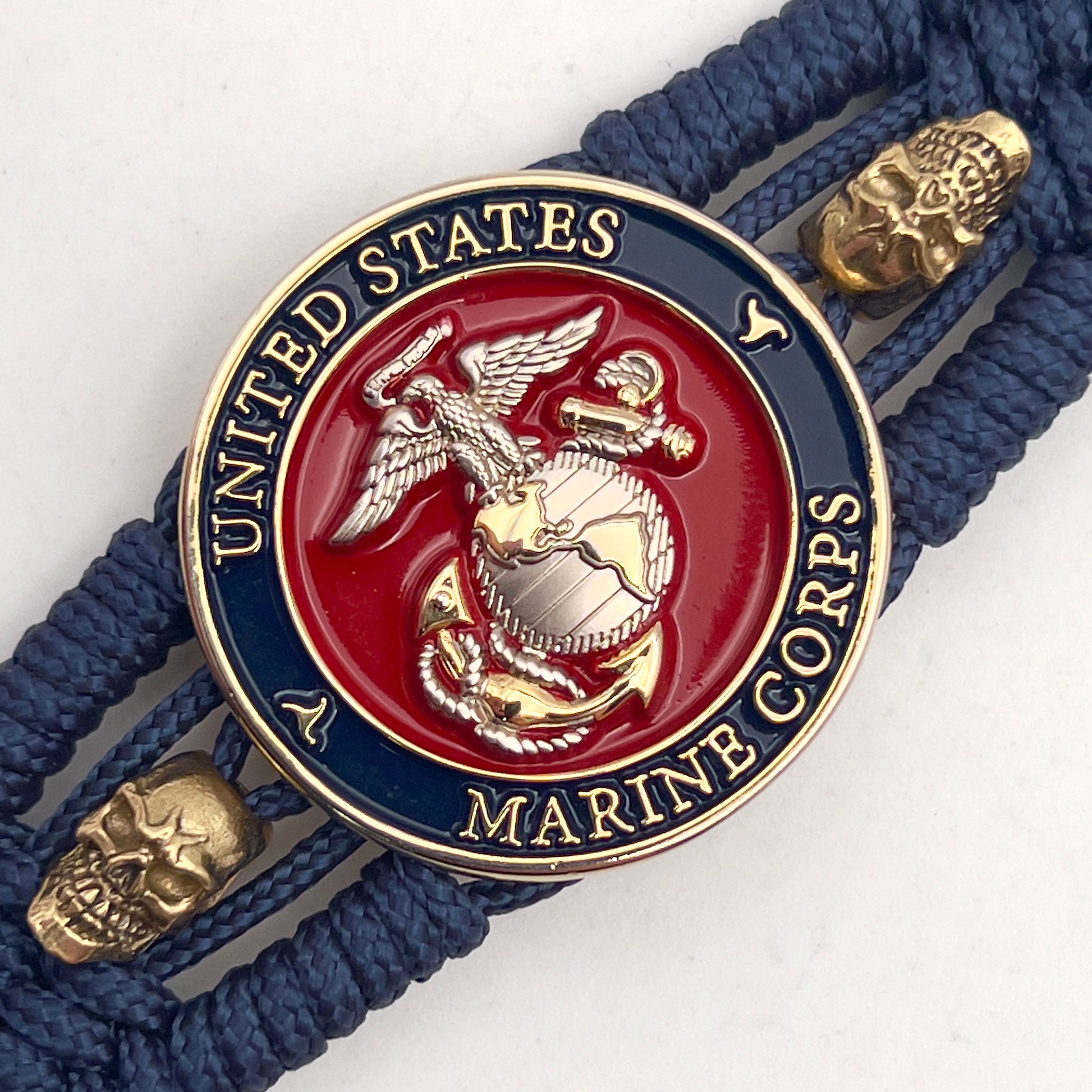 Thin, medium width, flexible and comfortable braided United States Marine Corps bracelet featuring a 24K gold and silver plated USMC officer Eagle Globe and Anchor medallion, two gold micro skulls, a gold Cobra style tactical buckle and woven with Navy blue type-1 paracord.