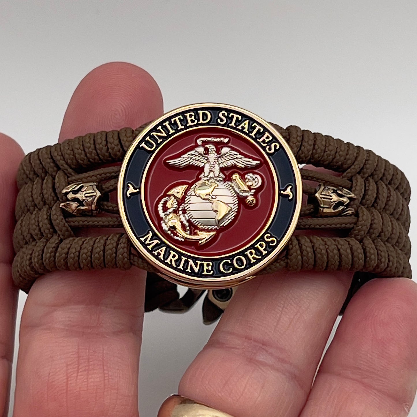 Thin, medium width, flexible and comfortable braided United States Marine Corps bracelet featuring a 24K gold and silver plated USMC officer Eagle Globe and Anchor medallion, two brass micro Spartan helmets, a gold Cobra style tactical buckle and woven with Coyote Brown type-1 paracord.