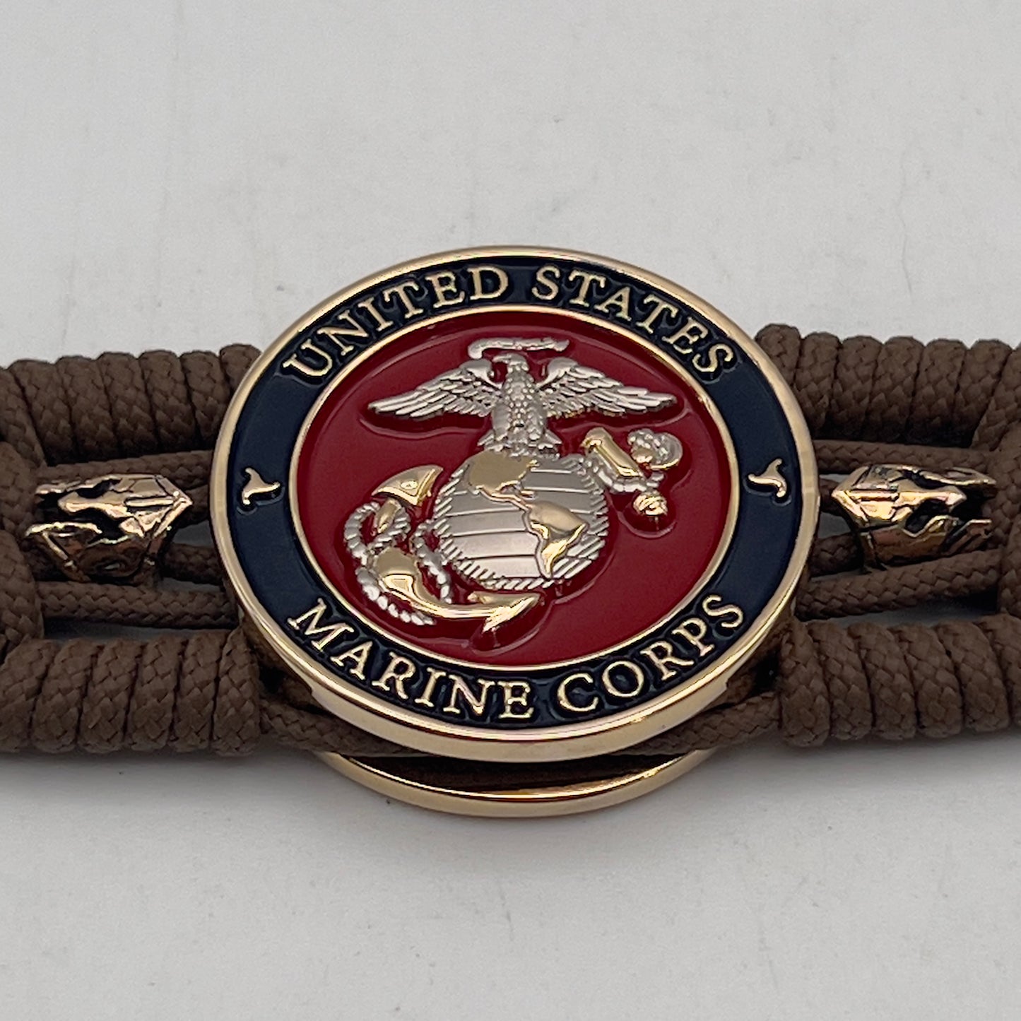Thin, medium width, flexible and comfortable braided United States Marine Corps bracelet featuring a 24K gold and silver plated USMC officer Eagle Globe and Anchor medallion, two brass micro Spartan helmets, a gold Cobra style tactical buckle and woven with Coyote Brown type-1 paracord.