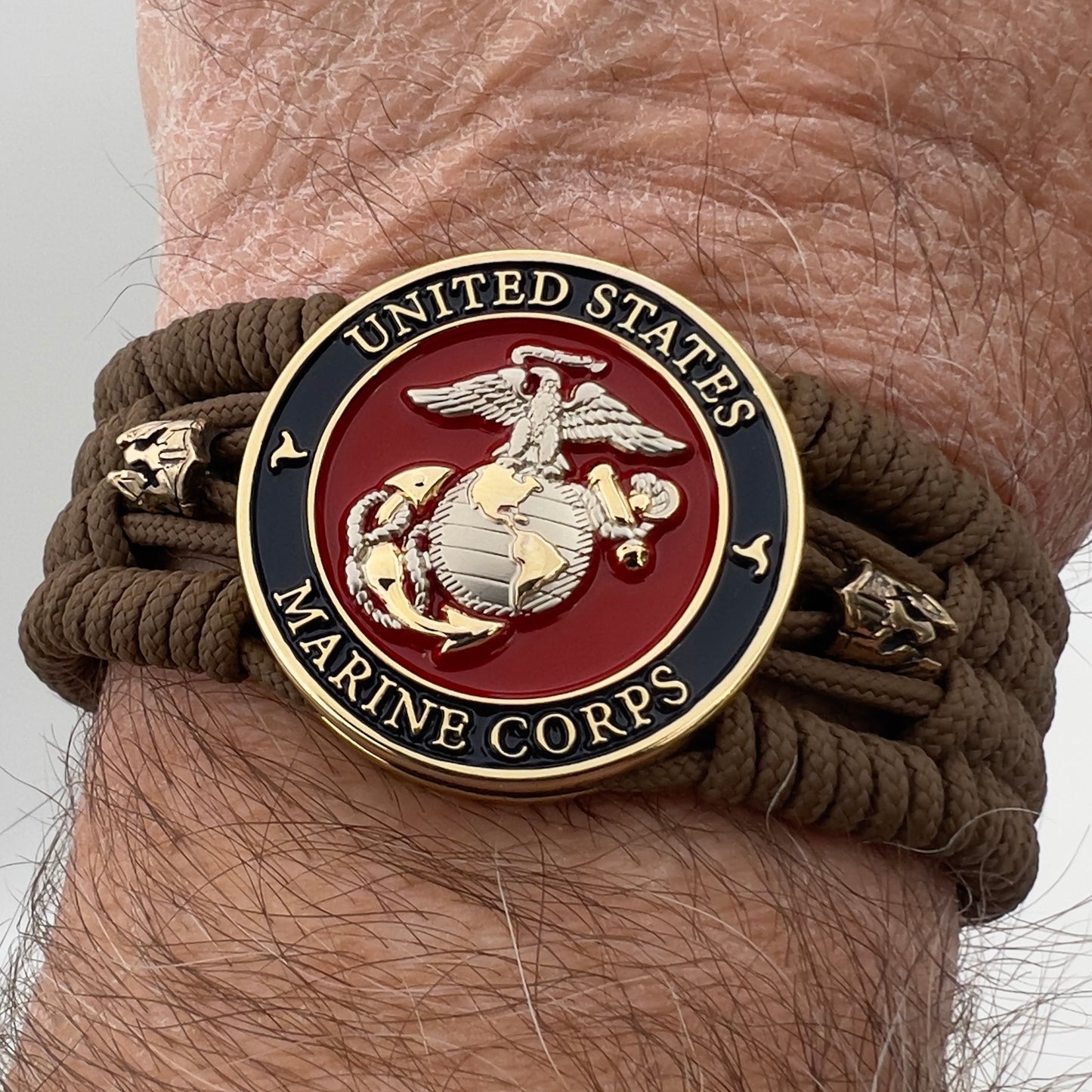 Thin, medium width, flexible and comfortable braided United States Marine Corps bracelet featuring a 24K gold and silver plated USMC officer Eagle Globe and Anchor medallion, two brass micro Spartan helmets, a gold Cobra style tactical buckle and woven with Coyote Brown type-1 paracord.