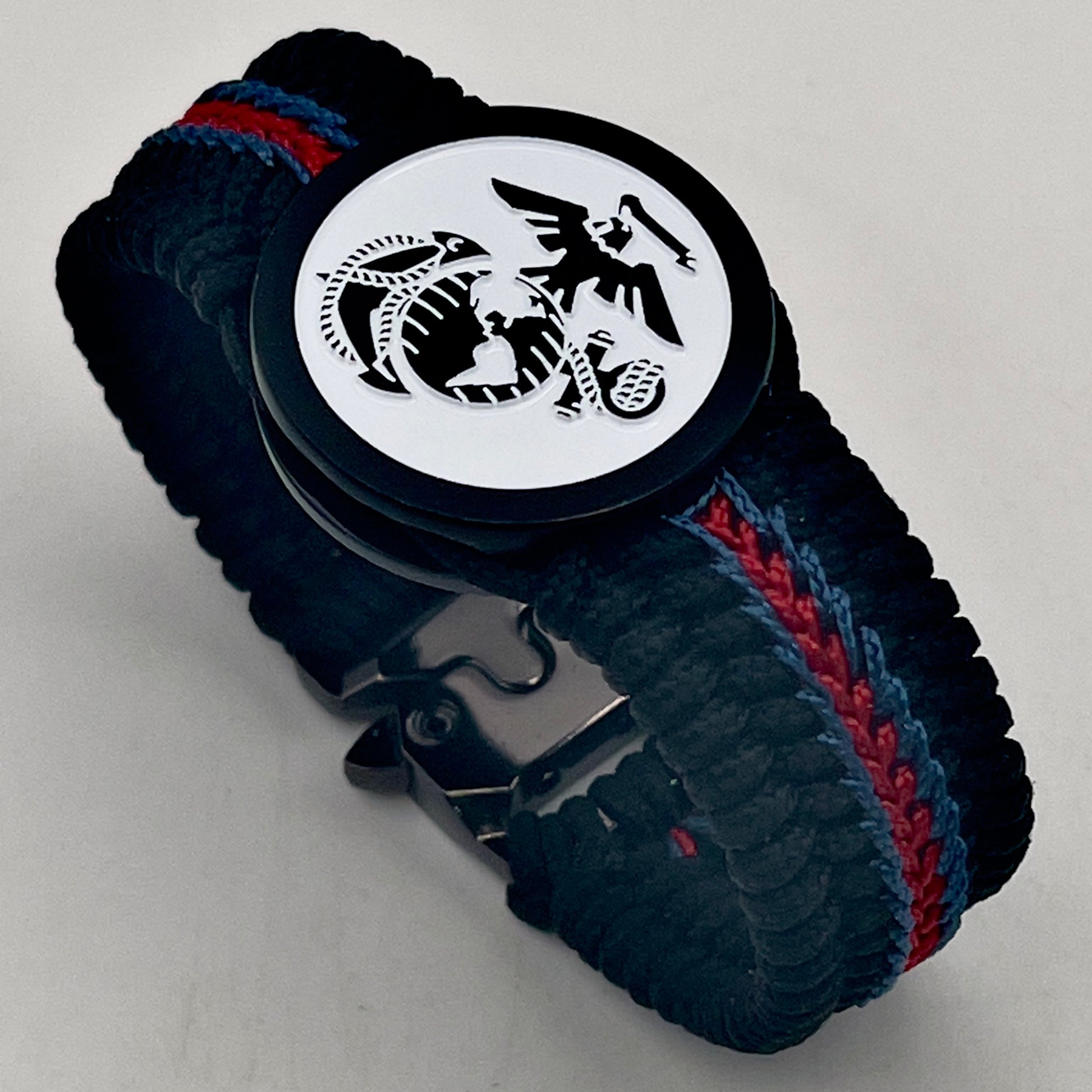 Thin, medium width, flexible and comfortable braided United States Marine Corps bracelet featuring a black USMC Eagle Globe and Anchor medallion, a black Cobra style tactical buckle and woven with black Type-1 paracord and stitched with red and blue nano cord.
