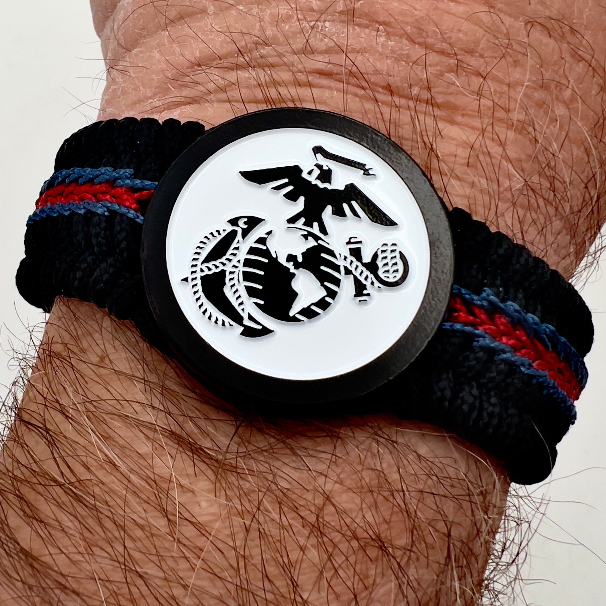 Thin, medium width, flexible and comfortable braided United States Marine Corps bracelet featuring a black USMC Eagle Globe and Anchor medallion, a black Cobra style tactical buckle and woven with black Type-1 paracord and stitched with red and blue nano cord.