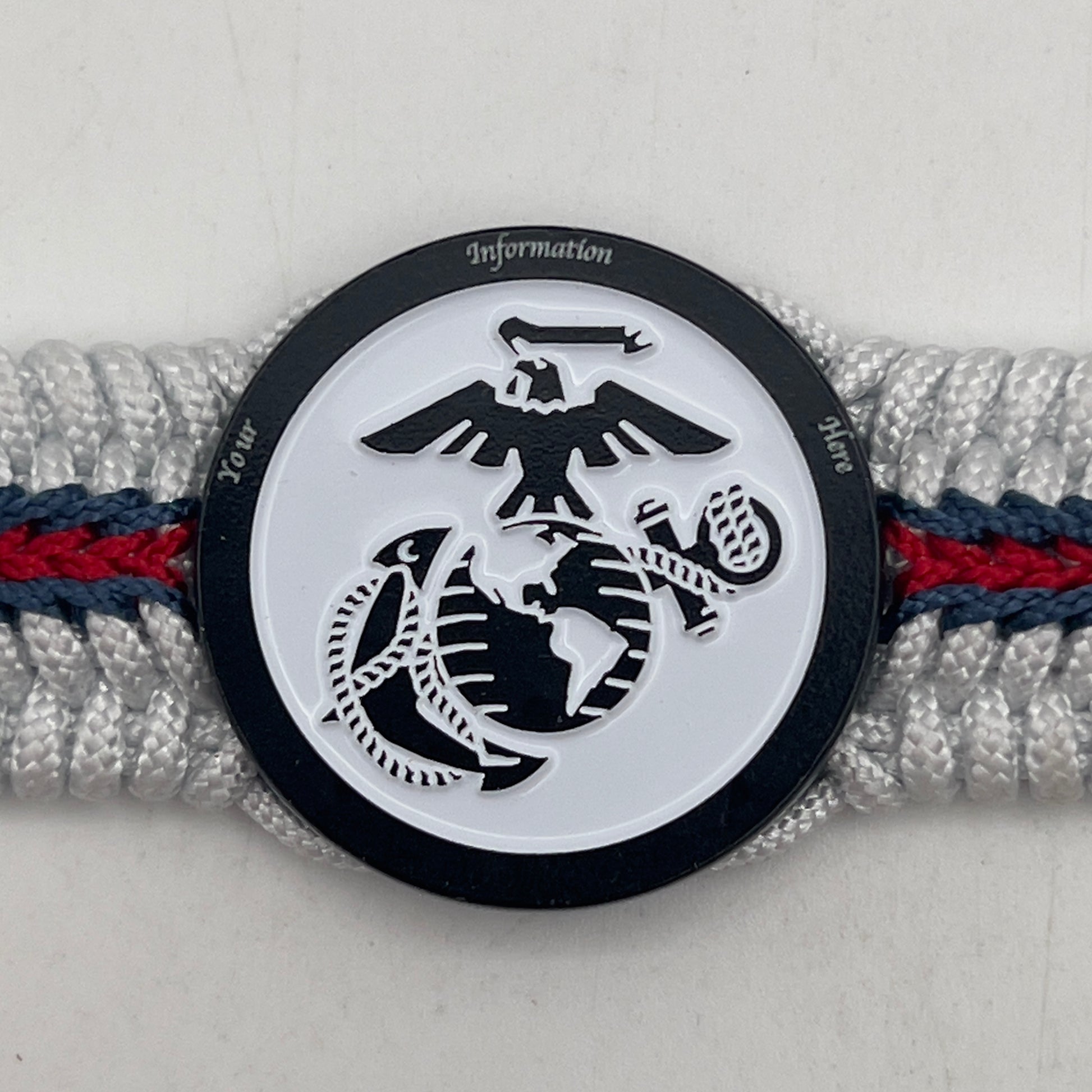Thin, medium width, flexible and comfortable braided United States Marine Corps bracelet featuring a black USMC Eagle Globe and Anchor medallion, a black Cobra style tactical buckle and woven with white Type-1 paracord and stitched with red and blue nano cord.