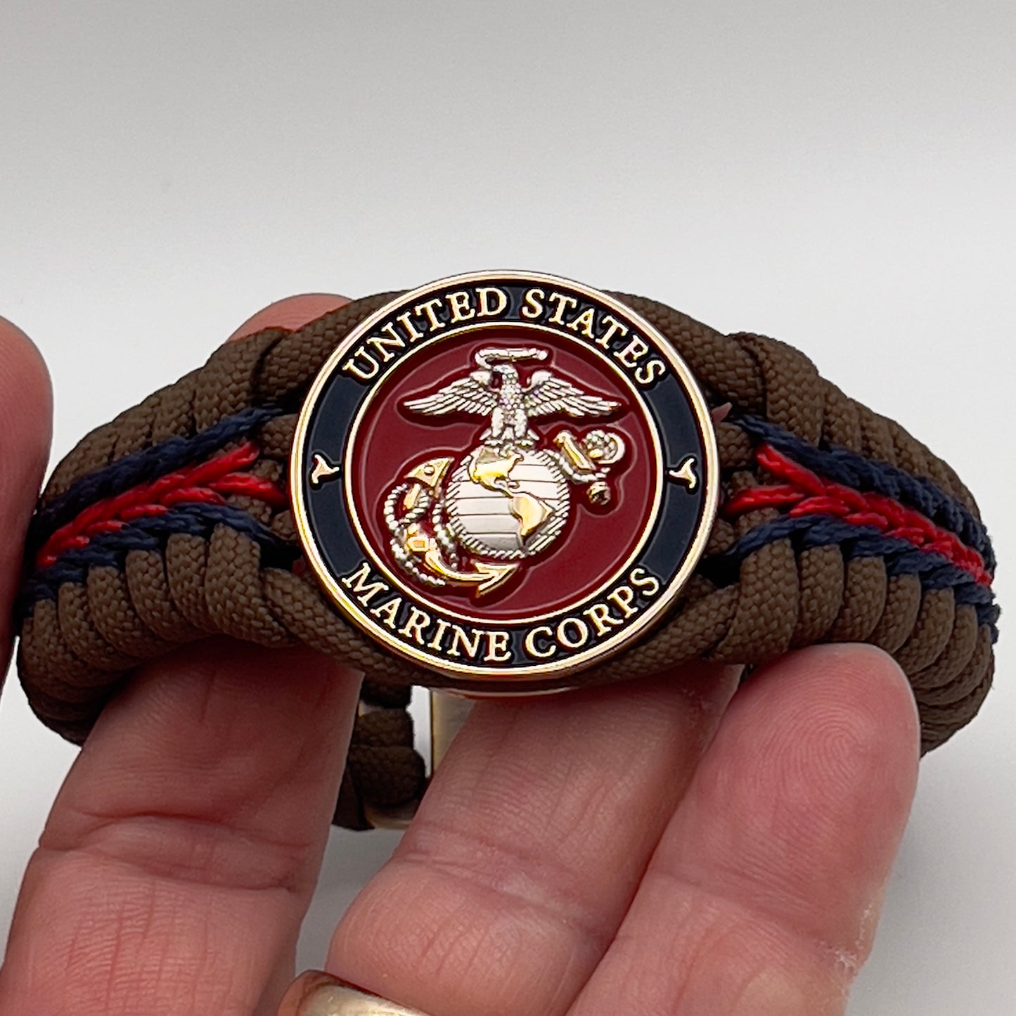 Thick, semi-rigid and comfortable braided United States Marine Corps bracelet featuring a 24K gold and silver plated USMC officer Eagle Globe and Anchor medallion, a Cobra style tactical buckle and woven with Coyote brown 550, Navy blue and Imperial red micro paracord.