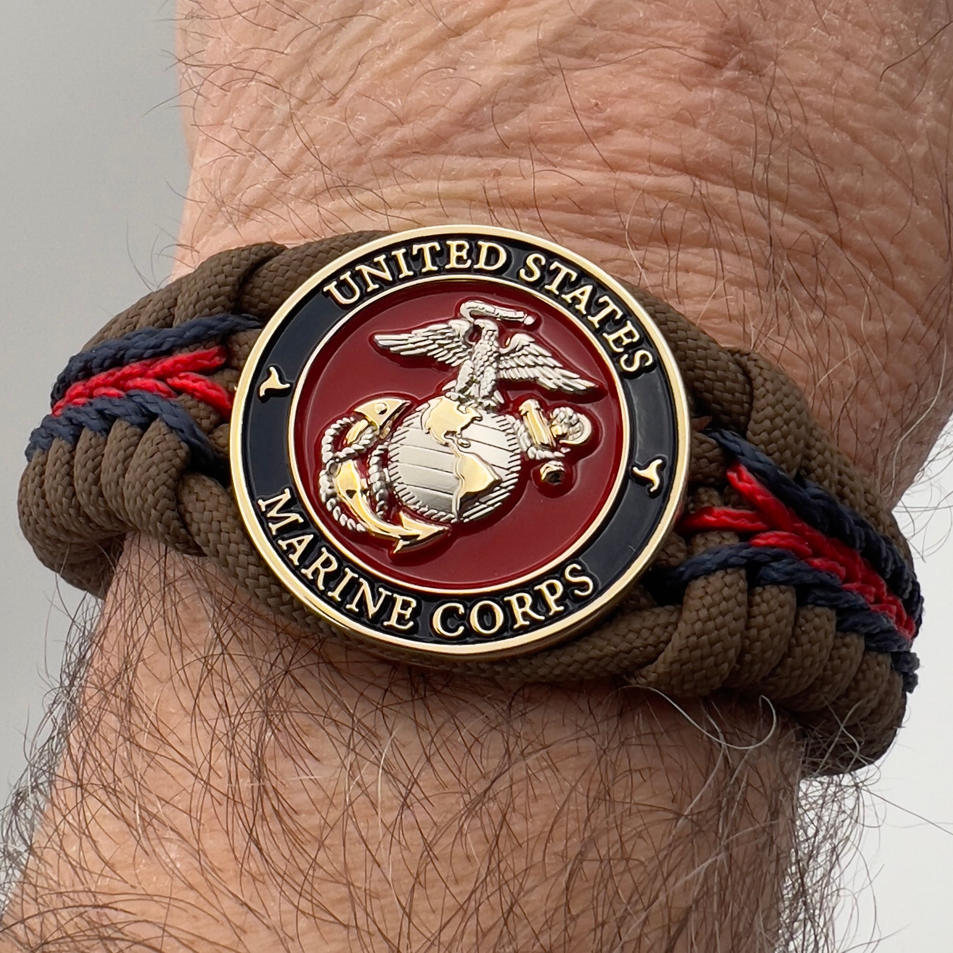 Thick, semi-rigid and comfortable braided United States Marine Corps bracelet featuring a 24K gold and silver plated USMC officer Eagle Globe and Anchor medallion, a Cobra style tactical buckle and woven with Coyote brown 550, Navy blue and Imperial red micro paracord.