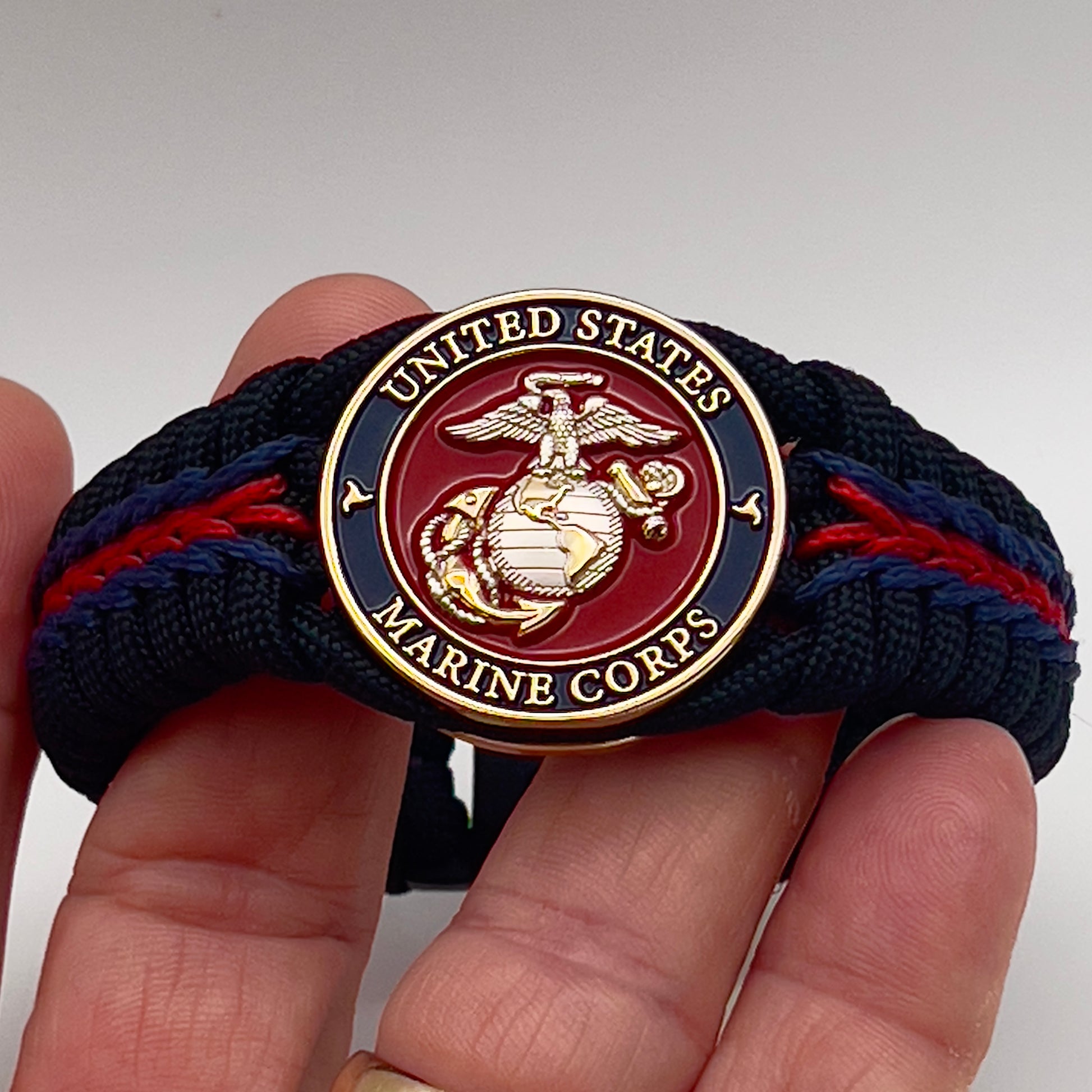 Thick, semi-rigid and comfortable braided United States Marine Corps bracelet featuring a 24K gold and silver plated USMC officer Eagle Globe and Anchor medallion, a black Cobra style tactical buckle and woven with Navy blue, Imperial red and black paracord.
