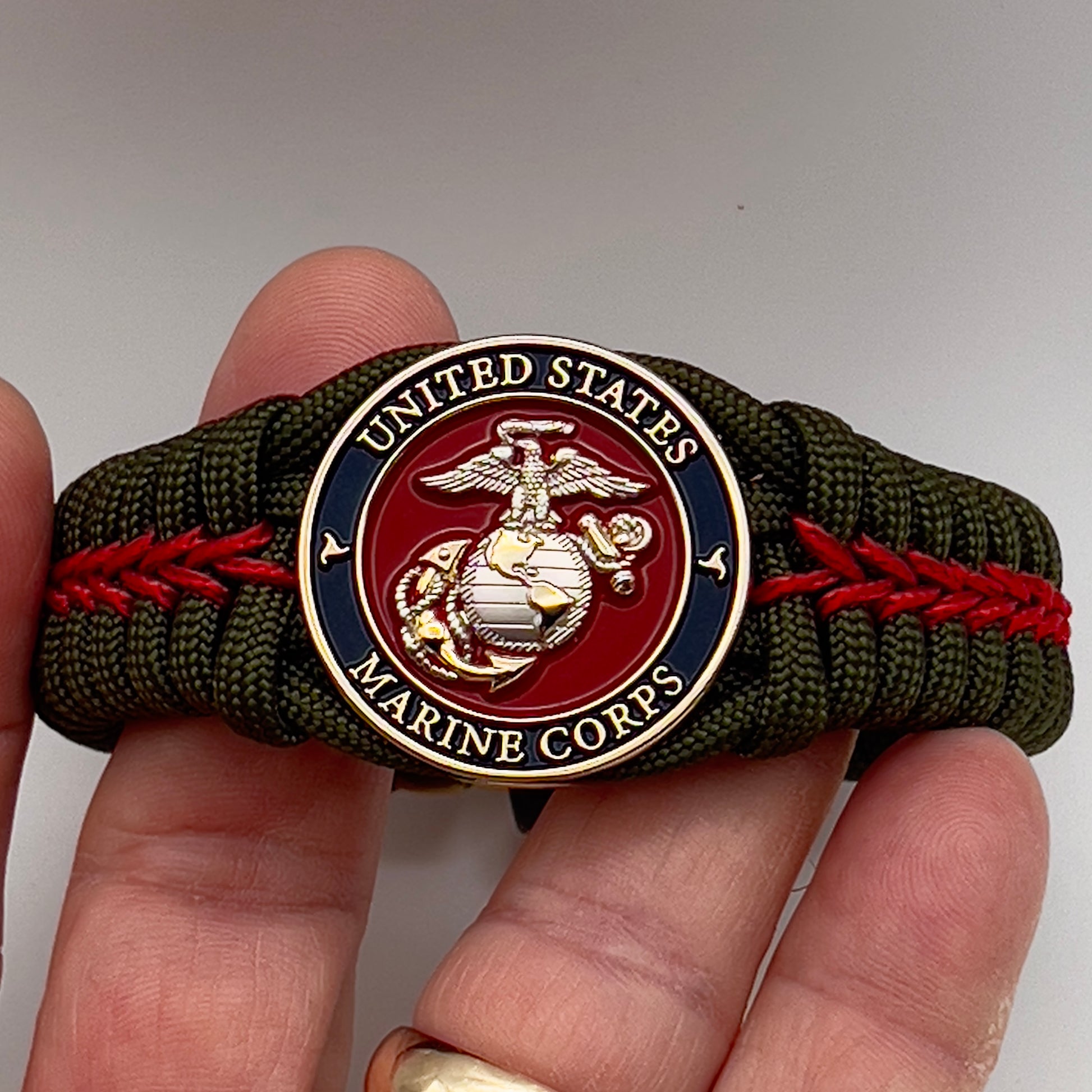 Thick, semi-rigid and comfortable braided United States Marine Corps bracelet featuring a 24K gold and silver plated USMC officer Eagle Globe and Anchor medallion, a gold Cobra style tactical buckle and woven with olive drab 550 and Imperial red micro paracord.