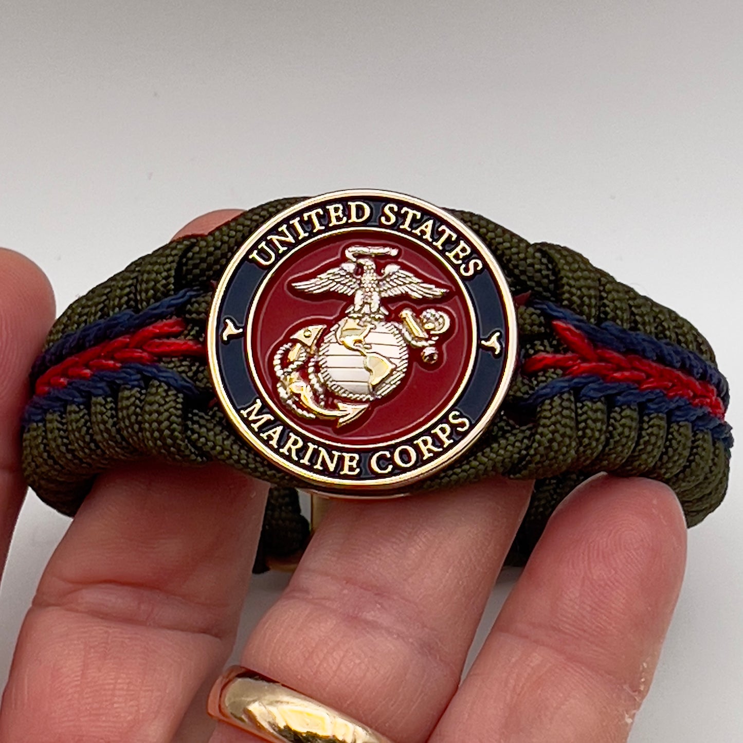 Thick, semi-rigid and comfortable braided United States Marine Corps bracelet featuring a 24K gold and silver plated USMC officer Eagle Globe and Anchor medallion, a gold Cobra style tactical buckle and woven with olive drab 550 and Imperial red and gold micro paracord.