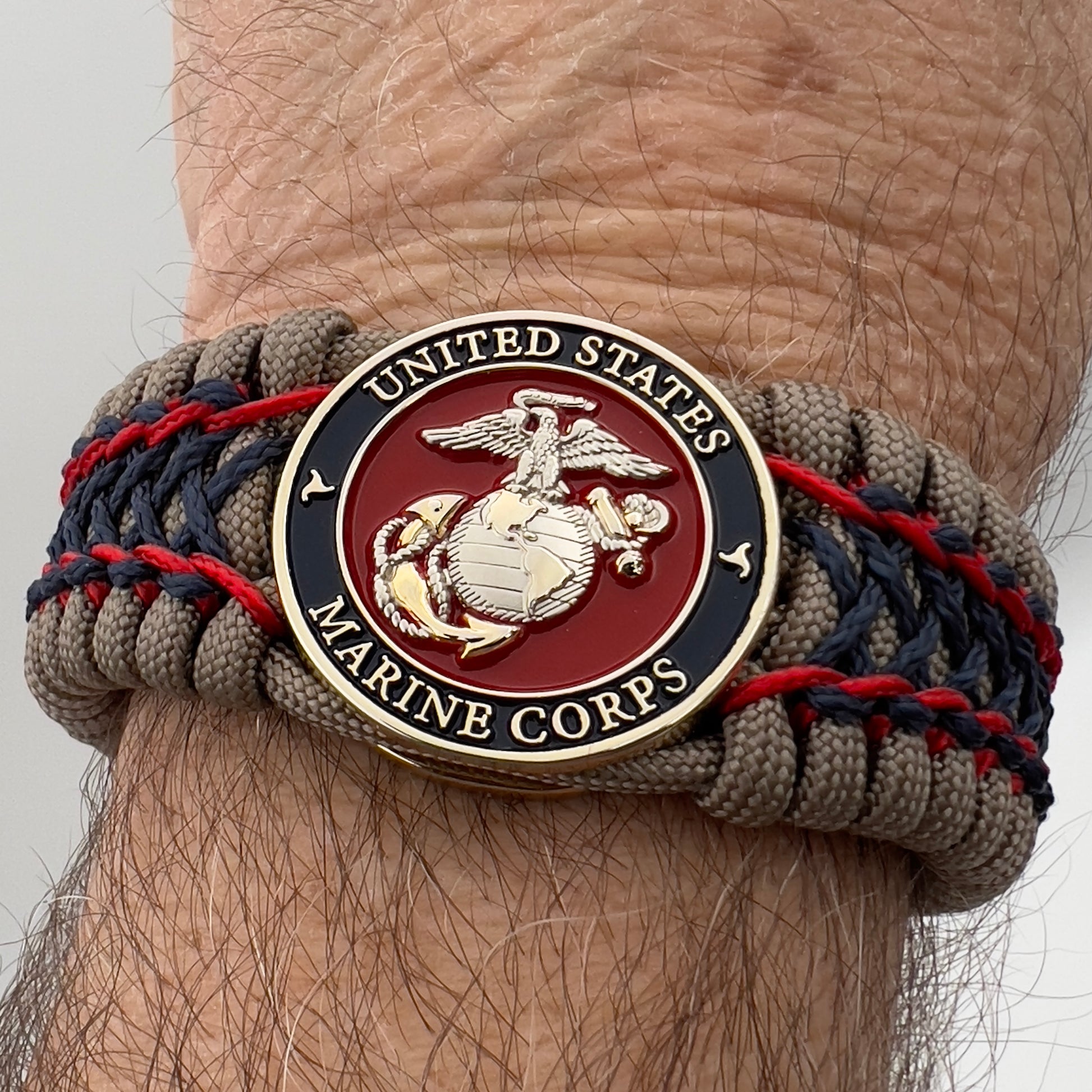 Thick, rigid and comfortable braided United States Marine Corps bracelet featuring a 24K gold and silver plated USMC officer Eagle Globe and Anchor medallion, a gold Cobra style tactical buckle and woven with tan 550 paracord and Imperial Red and Navy Blue micro cord.