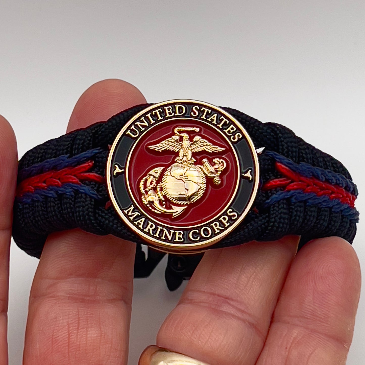 Thick, rigid and comfortable braided United States Marine Corps bracelet featuring a 24K gold plated USMC officer Eagle Globe and Anchor medallion, a black Cobra style tactical buckle and woven with black 550, Imperial red and Navy Blue micro paracord.