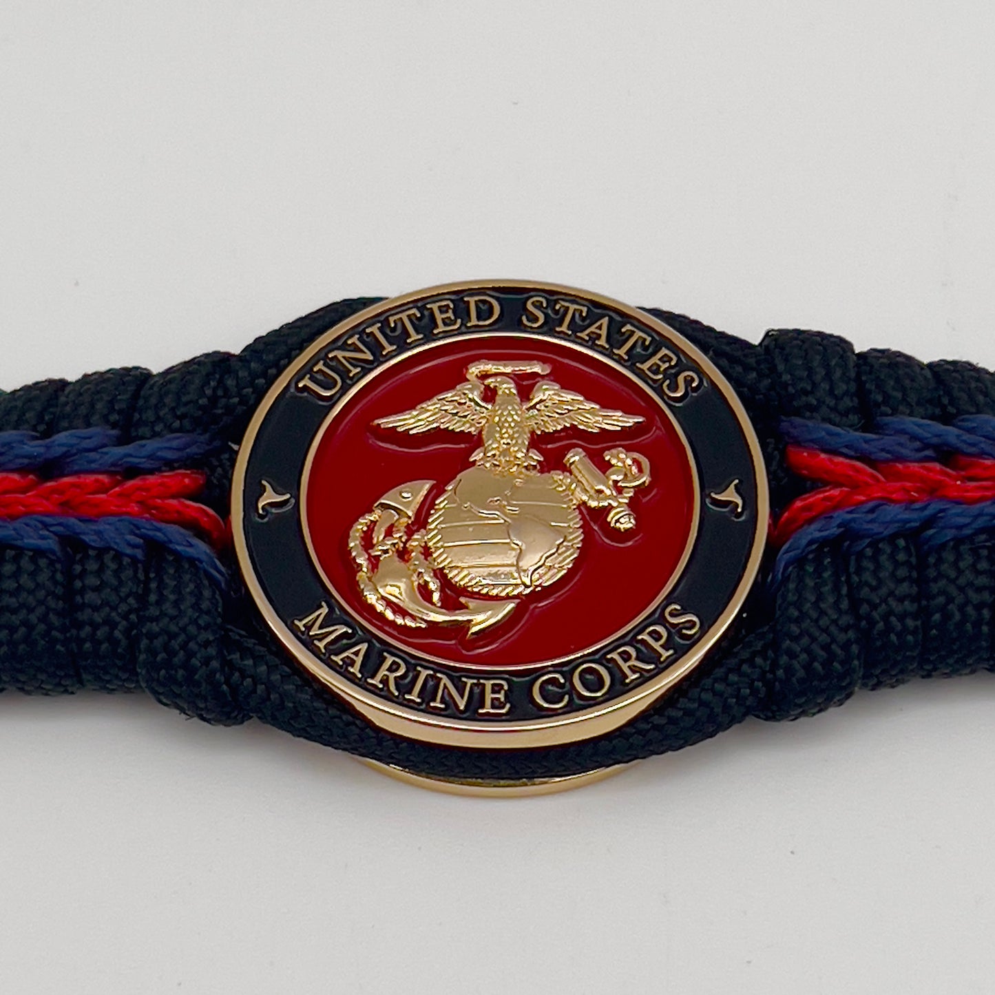 Thick, rigid and comfortable braided United States Marine Corps bracelet featuring a 24K gold plated USMC officer Eagle Globe and Anchor medallion, a black Cobra style tactical buckle and woven with black 550, Imperial red and Navy Blue micro paracord.