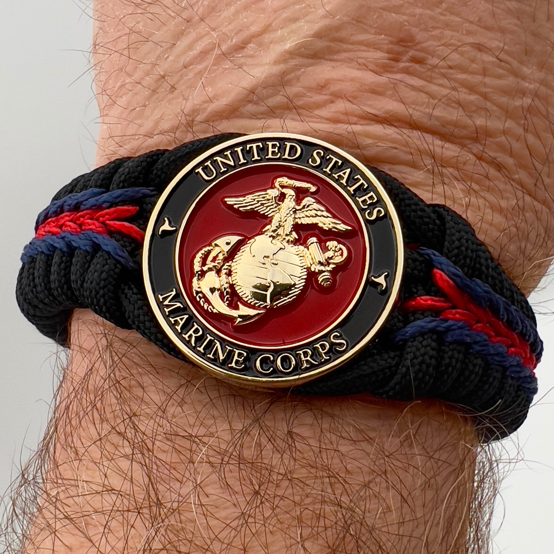 Thick, rigid and comfortable braided United States Marine Corps bracelet featuring a 24K gold plated USMC officer Eagle Globe and Anchor medallion, a black Cobra style tactical buckle and woven with black 550, Imperial red and Navy Blue micro paracord.