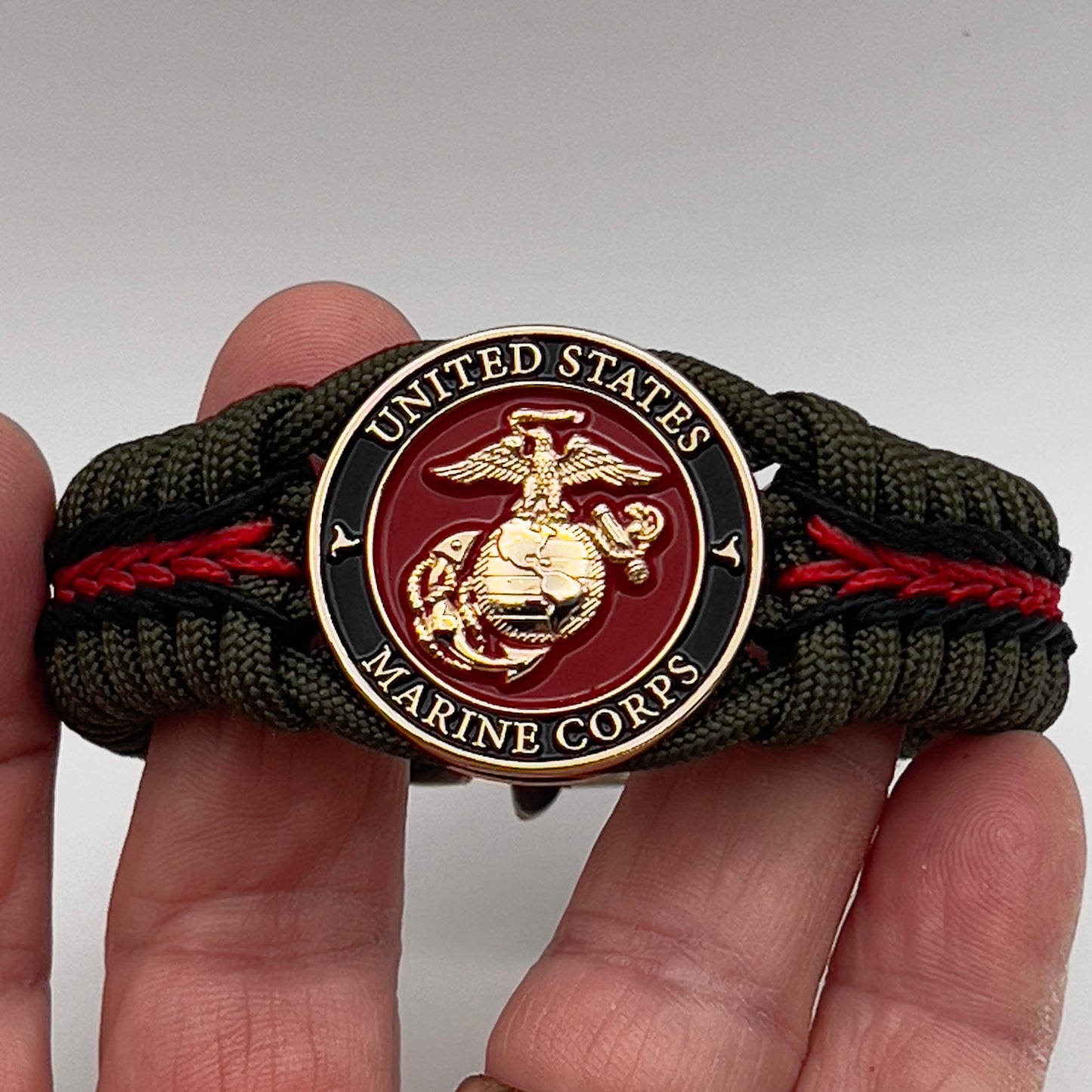 Thick, rigid and comfortable braided United States Marine Corps bracelet featuring a 24K gold plated USMC officer Eagle Globe and Anchor medallion, a gold Cobra style tactical buckle and woven with olive drab 550 paracord and black and Imperial red micro cord.