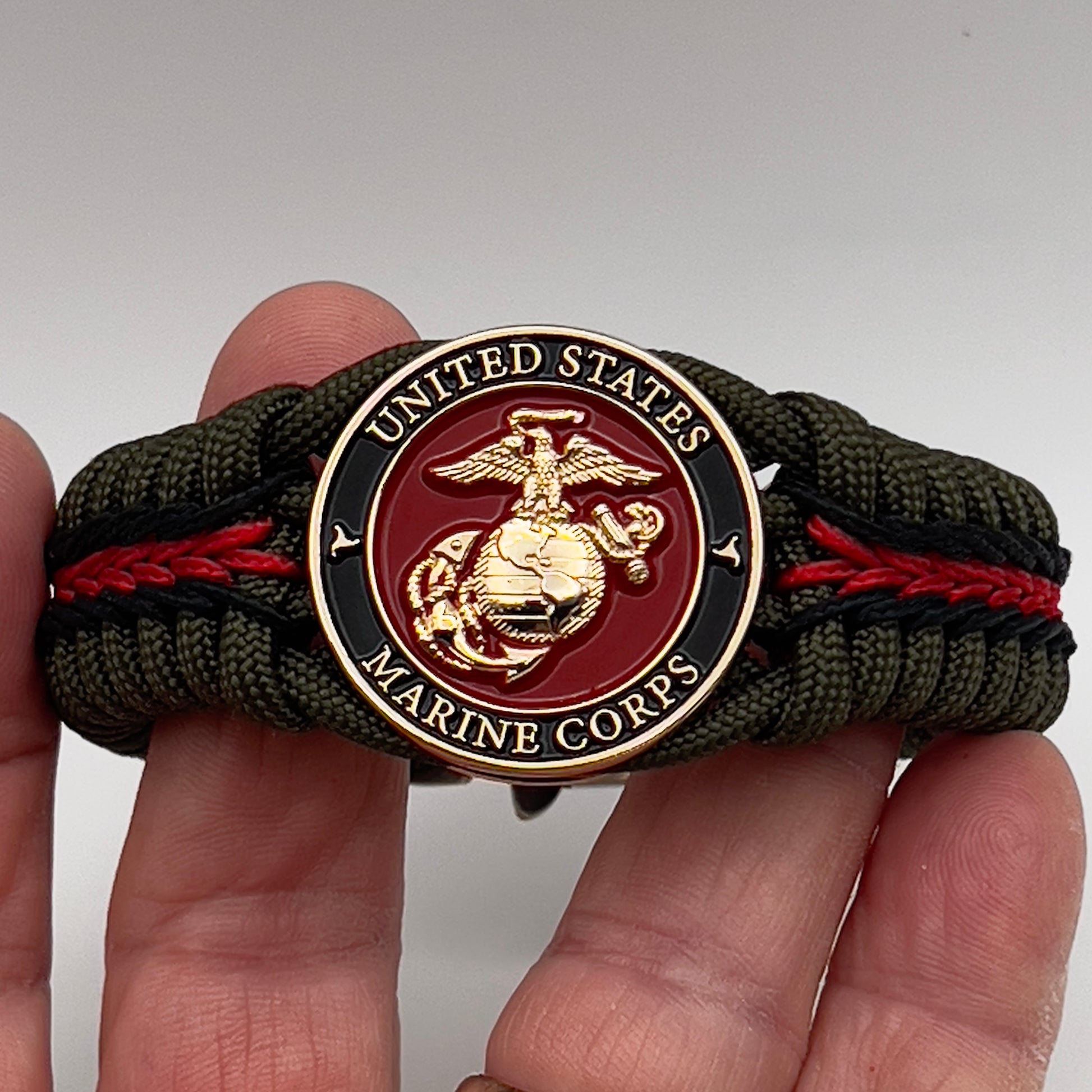 Thick, rigid and comfortable braided United States Marine Corps bracelet featuring a 24K gold plated USMC officer Eagle Globe and Anchor medallion, a gold Cobra style tactical buckle and woven with olive drab 550 paracord and black and Imperial red micro cord.