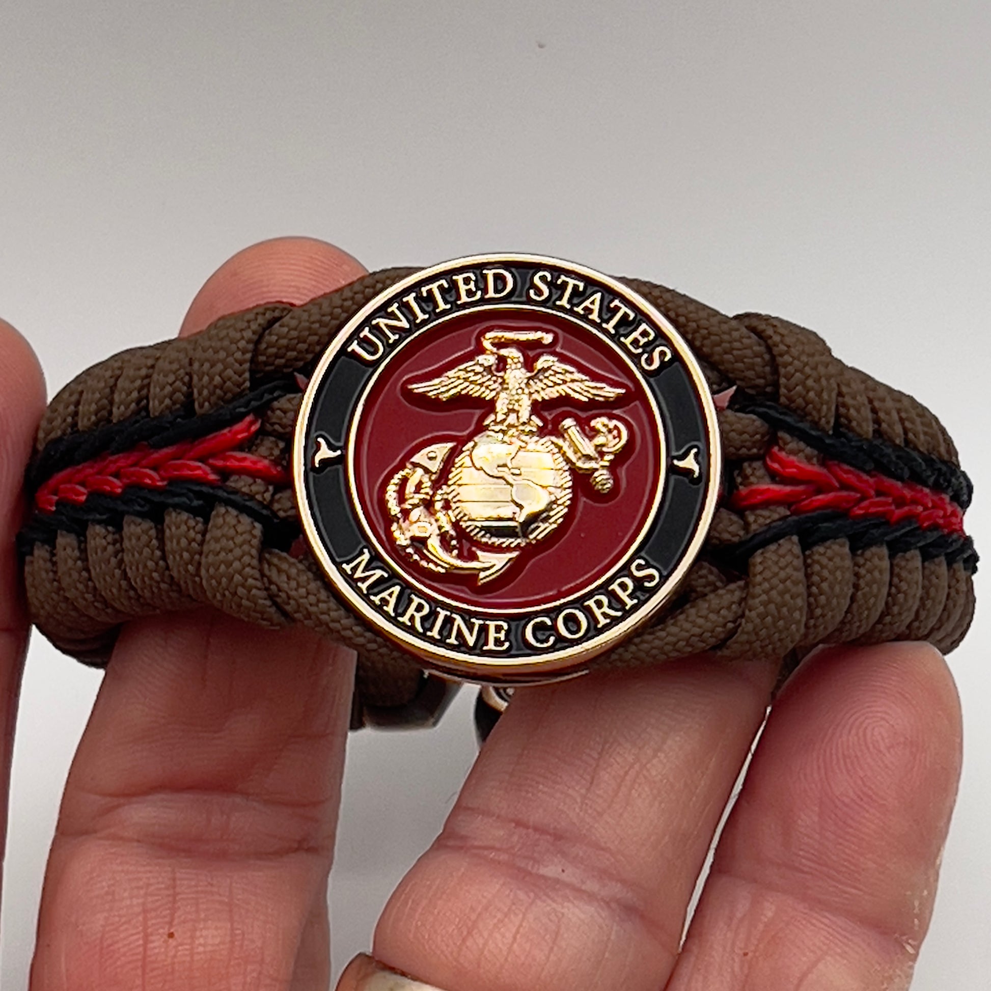 Thick, rigid and comfortable braided United States Marine Corps bracelet featuring a 24K gold plated USMC Eagle Globe and Anchor medallion, a gold Cobra style tactical buckle and woven with Coyote Brown 550 paracord and Imperial Red and black micro cord.
