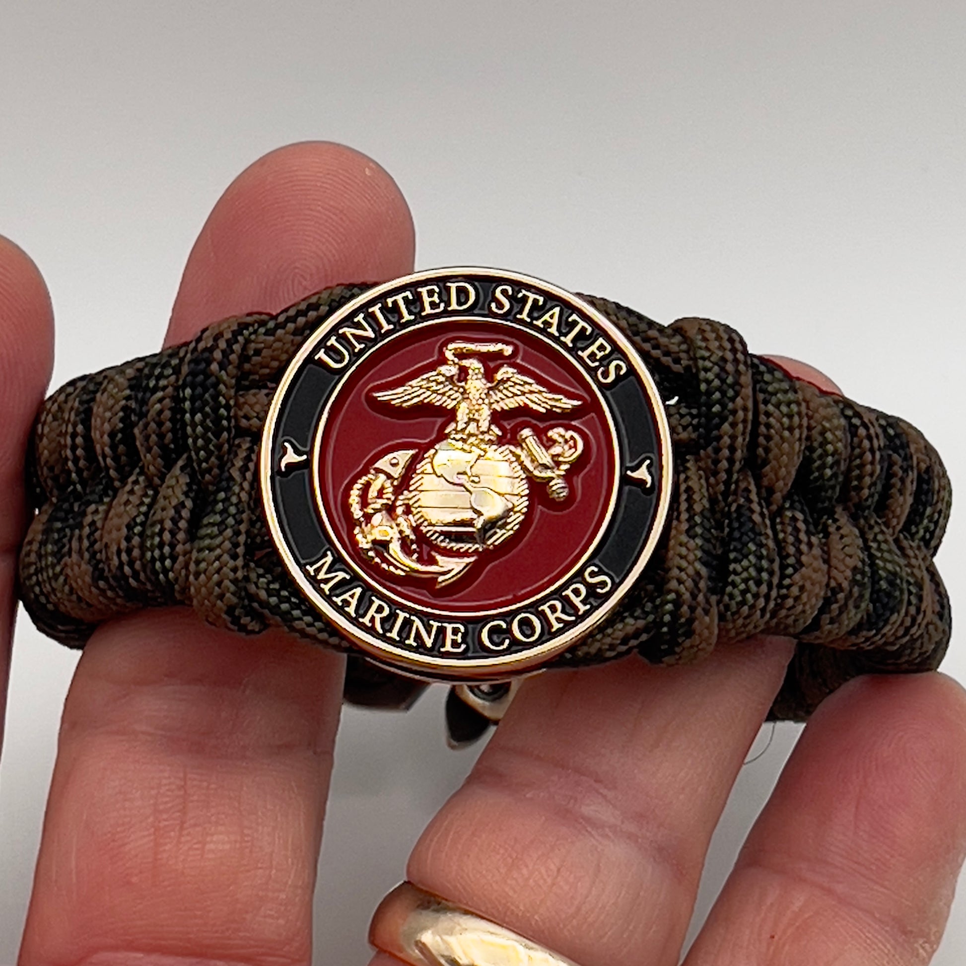 Thick, semi-rigid and comfortable braided United States Marine Corps bracelet featuring a 24K gold plated USMC Eagle Globe and Anchor medallion, a gold Cobra style tactical buckle and woven with Woodland camo 550 paracord.