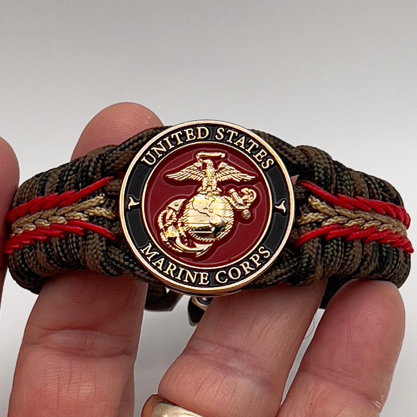 Thick, rigid and comfortable braided United States Marine Corps bracelet featuring a 24K gold plated USMC Eagle Globe and Anchor medallion, a gold Cobra style tactical buckle and woven with Woodland camo 550 paracord and Imperial Red and Gold micro cord.
