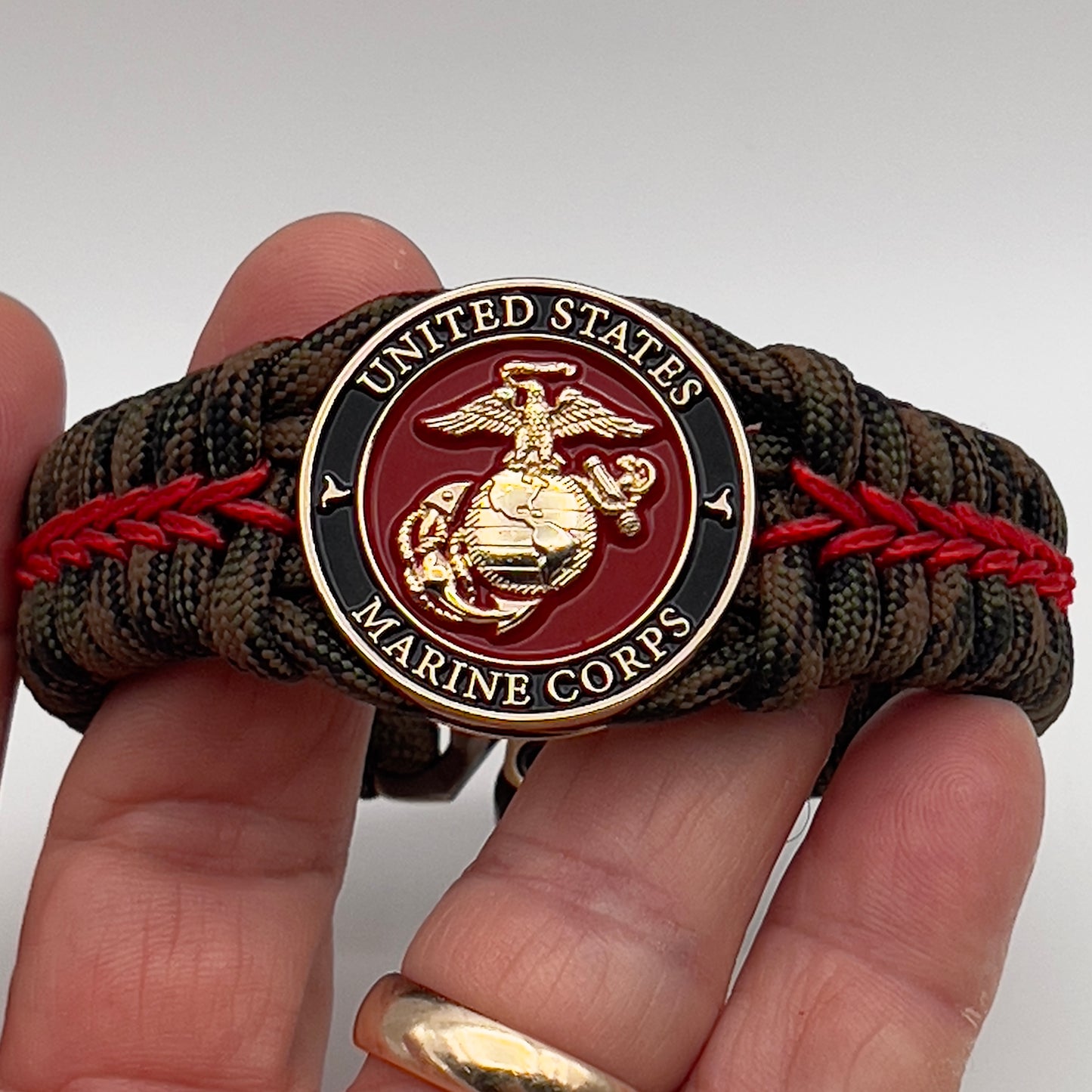 Thick, rigid and comfortable braided United States Marine Corps bracelet featuring a 24K gold plated USMC Eagle Globe and Anchor medallion, a gold Cobra style tactical buckle and woven with Woodland Camo 550 paracord and Imperial Red micro cord.