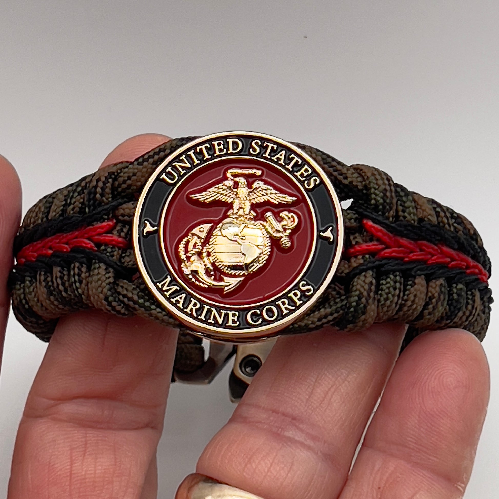 Thick, semi-rigid and comfortable braided United States Marine Corps bracelet featuring a 24K gold plated USMC Eagle Globe and Anchor medallion, a gold Cobra style tactical buckle and woven with Woodland camo 550 paracord and black and Imperial red micro cord.