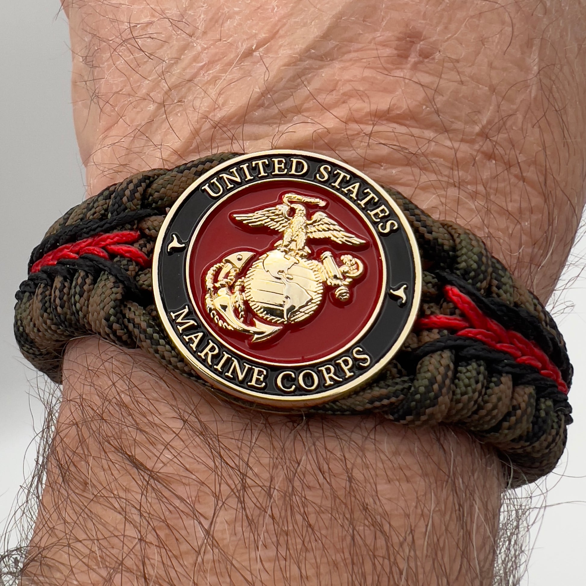 Thick, semi-rigid and comfortable braided United States Marine Corps bracelet featuring a 24K gold plated USMC Eagle Globe and Anchor medallion, a gold Cobra style tactical buckle and woven with Woodland camo 550 paracord and black and Imperial red micro cord.