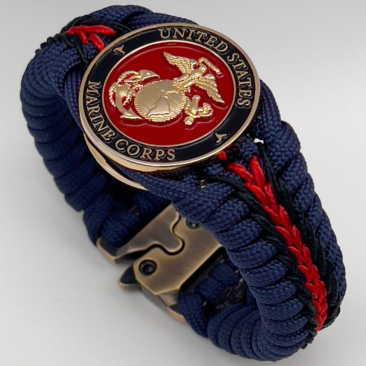 Thick, rigid and comfortable braided United States Marine Corps bracelet featuring a 24K gold plated USMC Eagle Globe and Anchor medallion, a gold Cobra style tactical buckle and woven with Navy Blue 550 paracord and Imperial Red and Black micro cord.