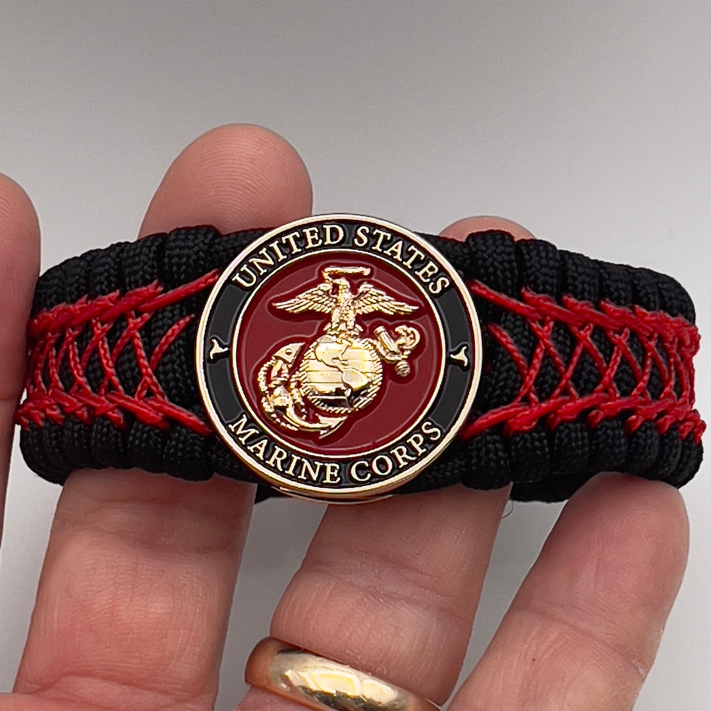 Thick, wide, rigid and comfortable braided United States Marine Corps bracelet featuring a 24K gold plated USMC Eagle Globe and Anchor medallion, a black Cobra style tactical buckle and woven with black 550 paracord and Imperial Red nano and micro cord.