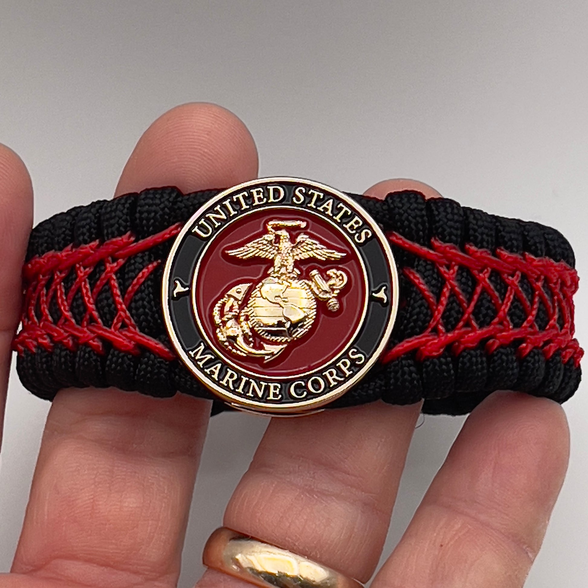 Thick, wide, rigid and comfortable braided United States Marine Corps bracelet featuring a 24K gold plated USMC Eagle Globe and Anchor medallion, a black Cobra style tactical buckle and woven with black 550 paracord and Imperial Red nano and micro cord.