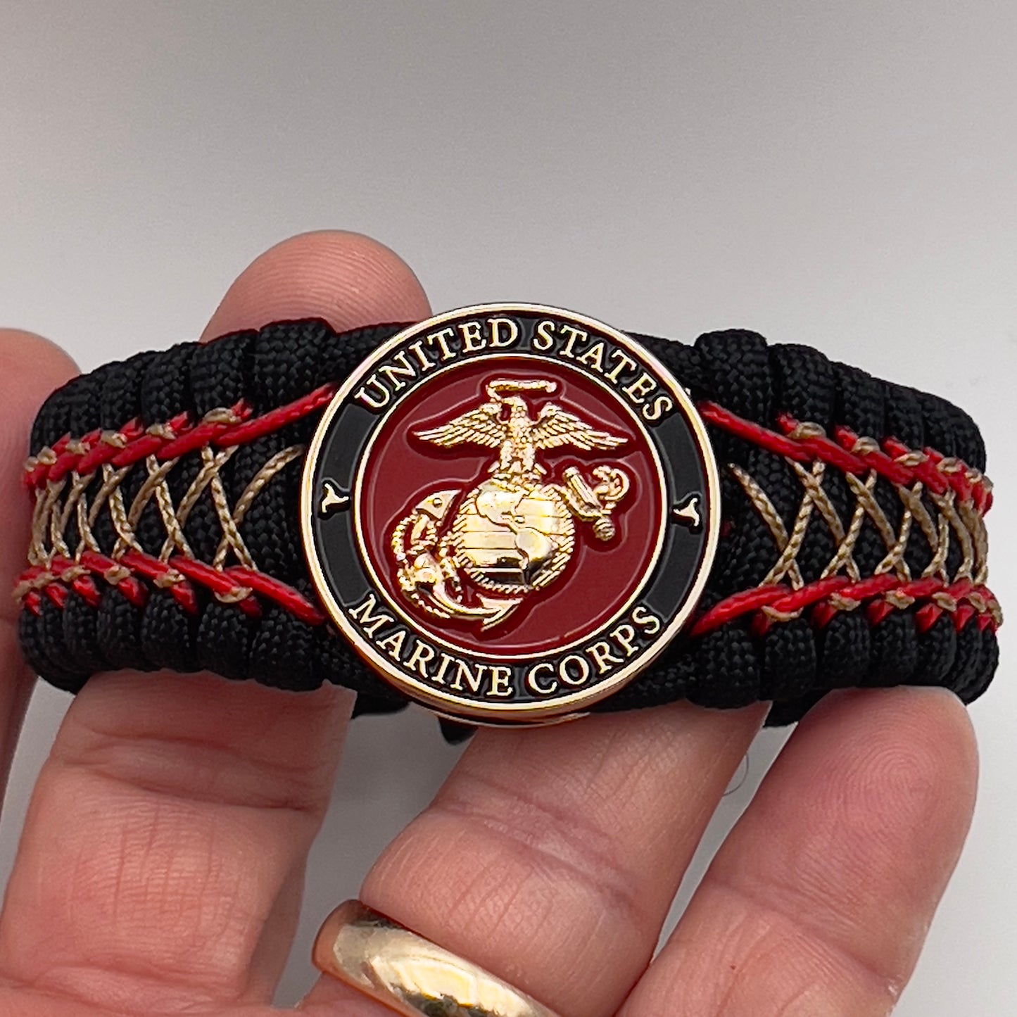 Thick, wide, rigid and comfortable braided United States Marine Corps bracelet featuring a 24K gold plated USMC Eagle Globe and Anchor medallion, a black Cobra style tactical buckle and woven with black 550 paracord and Imperial Red micro and gold nano cord.