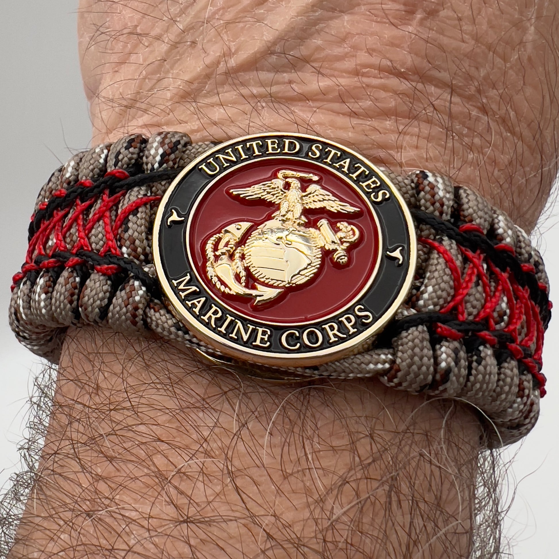 Thick, wide, rigid and comfortable braided United States Marine Corps bracelet featuring a 24K gold plated USMC Eagle Globe and Anchor medallion, a gold Cobra style tactical buckle and woven with Desert Camo 550 paracord, black micro cord and Imperial Red nano cord.