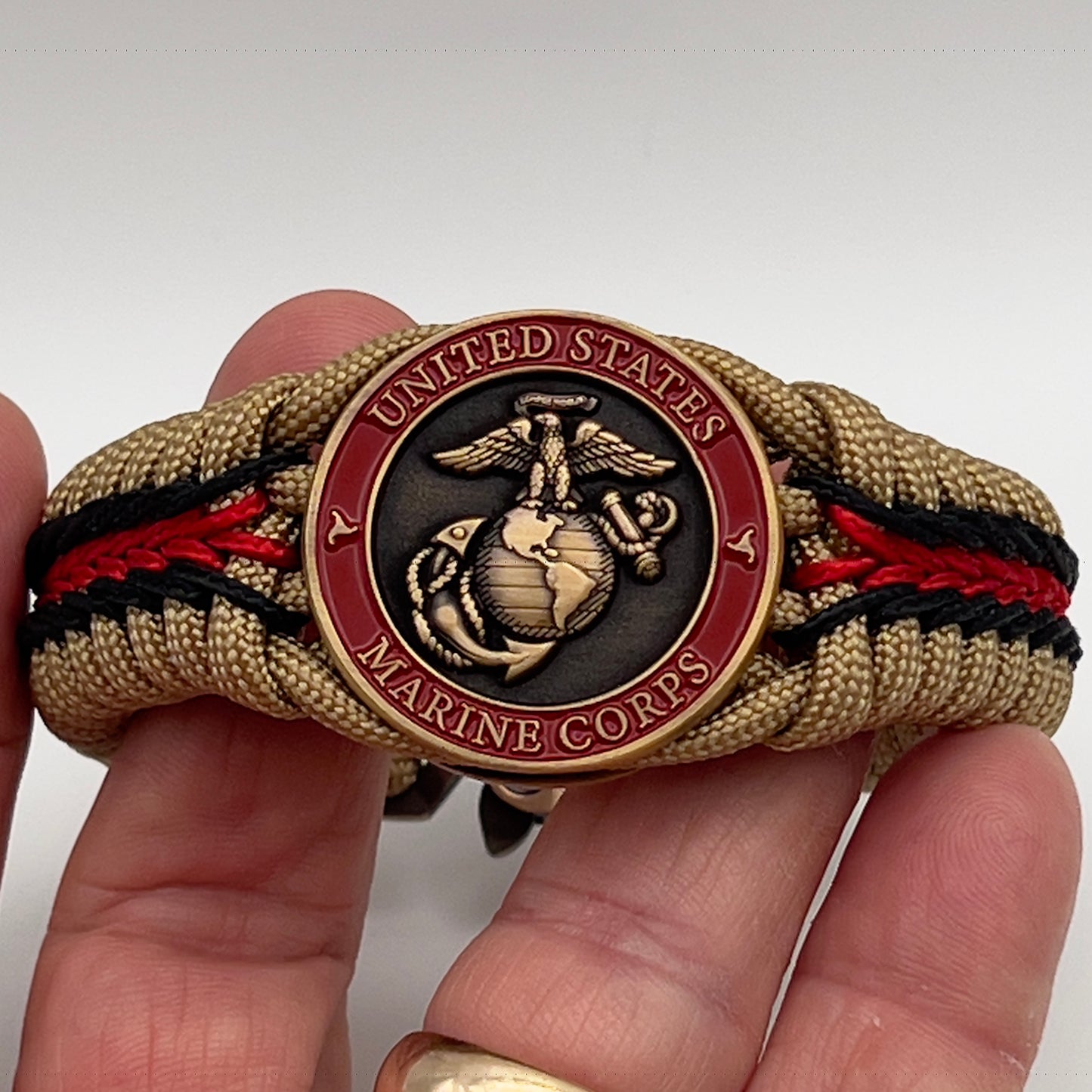 Thick, medium width, rigid and comfortable braided United States Marine Corps bracelet featuring a 24K antique gold plated USMC Eagle Globe and Anchor medallion, a gold Cobra style tactical buckle and woven with gold 550 paracord and Imperial red and black micro cord.