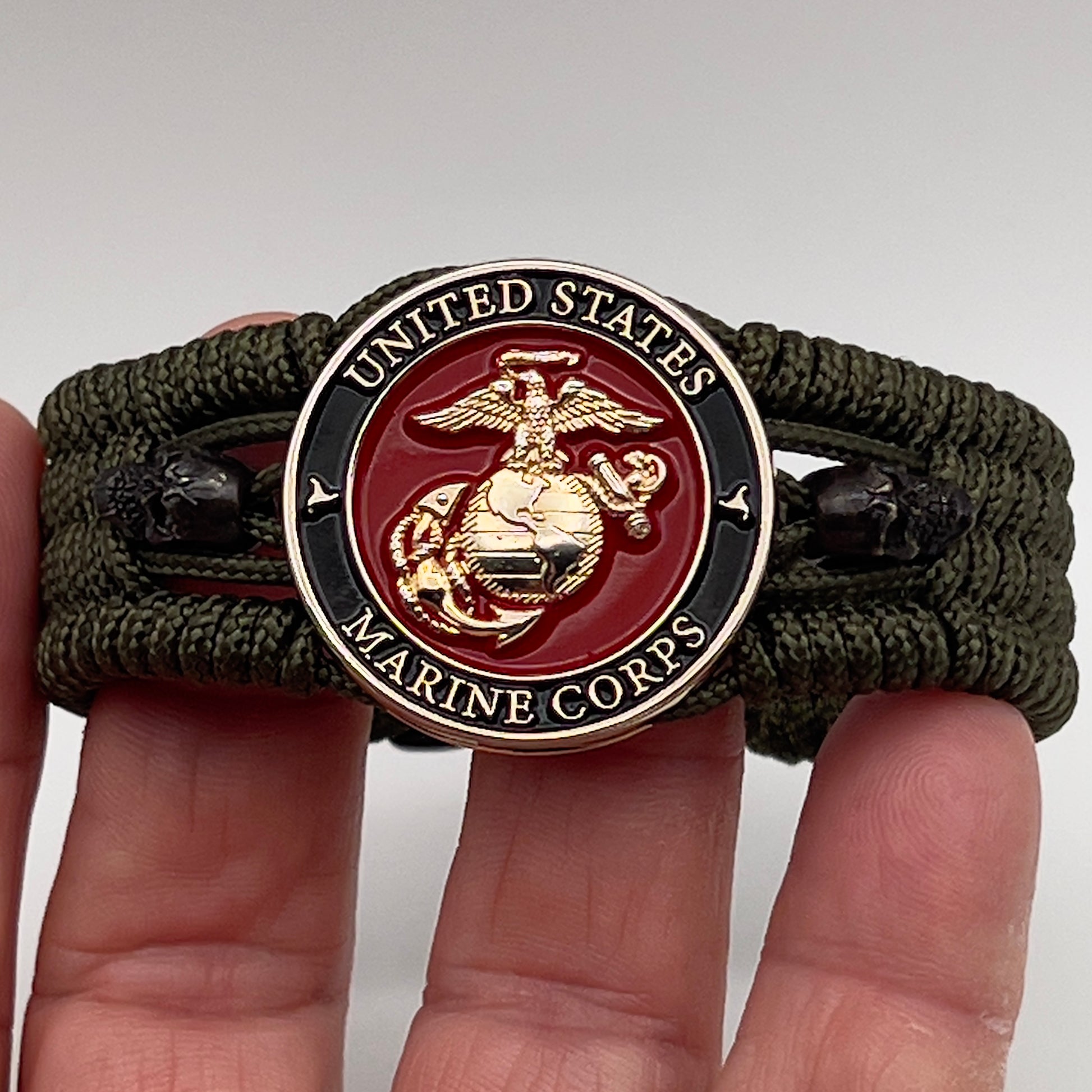 Thin, medium width, flexible and comfortable braided United States Marine Corps bracelet featuring a 24K gold plated USMC officer Eagle Globe and Anchor medallion, two black micro skulls, a black Cobra style tactical buckle and woven with Olive Drab Type-1 paracord.