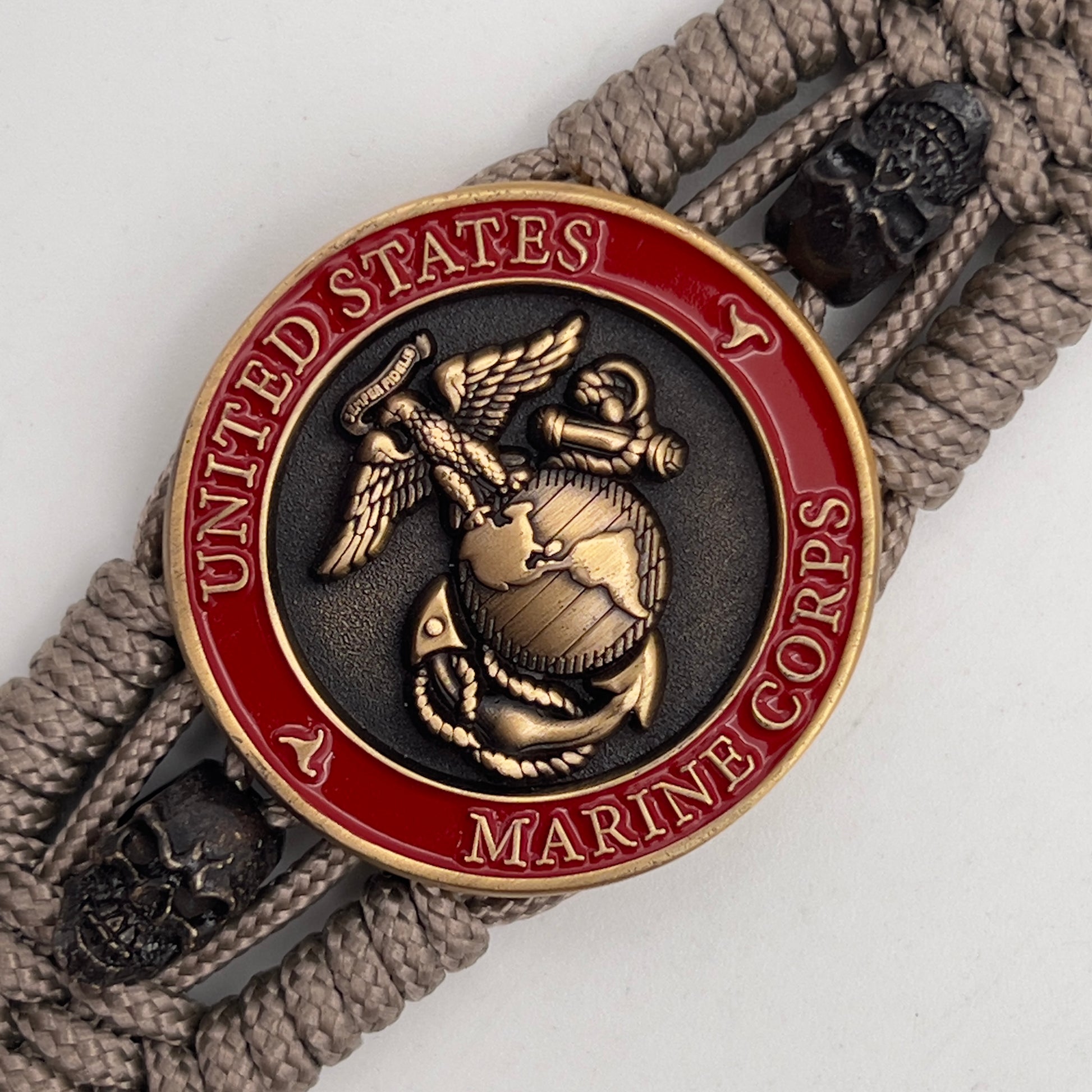 Thin, medium width, flexible and comfortable braided United States Marine Corps bracelet featuring a 24K antique gold plated USMC officer Eagle Globe and Anchor medallion, two black skulls, a black Cobra style tactical buckle and woven with tan Type-1 paracord.