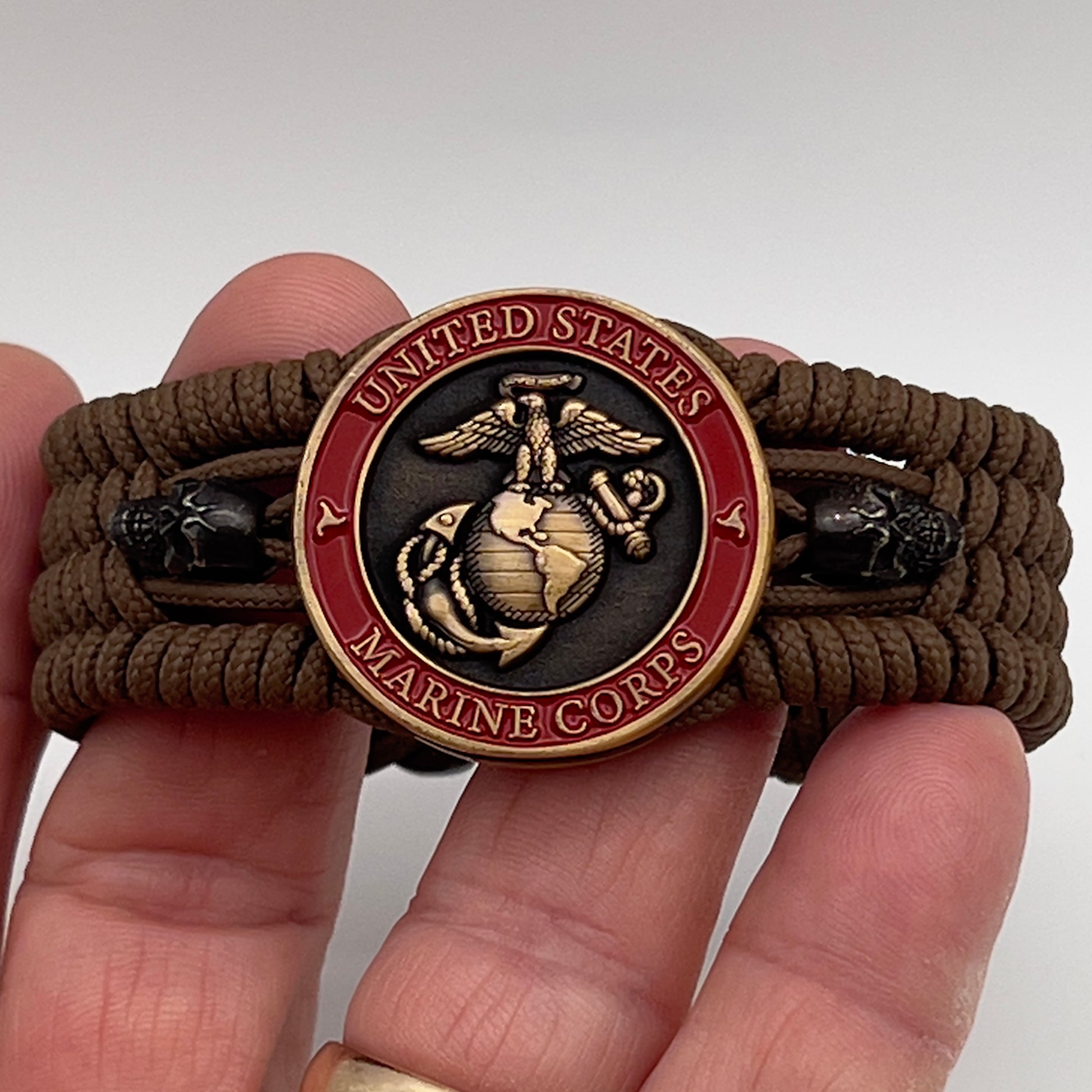 Thin, medium width, flexible and comfortable braided United States Marine Corps bracelet featuring a 24K antique gold plated USMC officer Eagle Globe and Anchor medallion, two black skulls, a black Cobra style tactical buckle and woven with Coyote Brown Type-1 paracord.