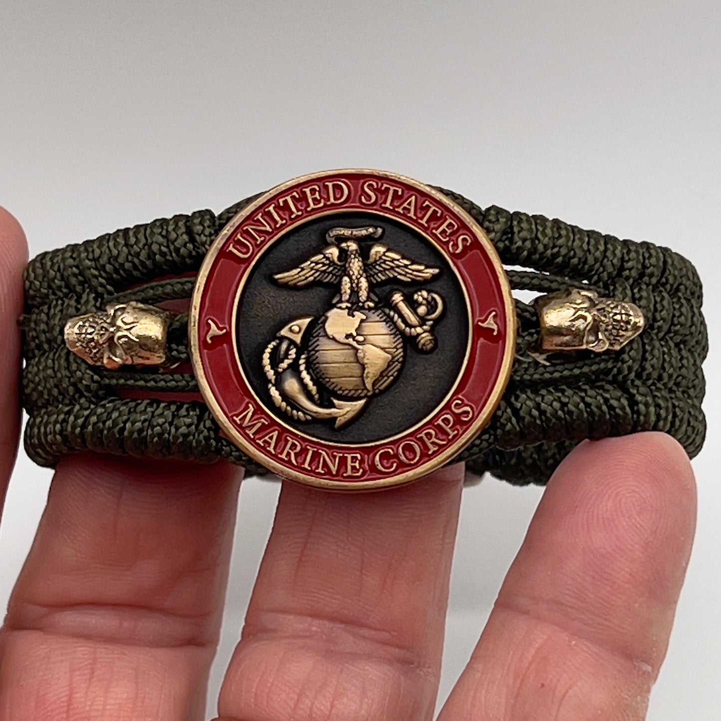 Thin, medium width, flexible and comfortable braided United States Marine Corps bracelet featuring a 24K antique gold plated USMC officer Eagle Globe and Anchor medallion, two gold plated skulls, a gold Cobra style tactical buckle and woven with Olive Drab Type-1 paracord.