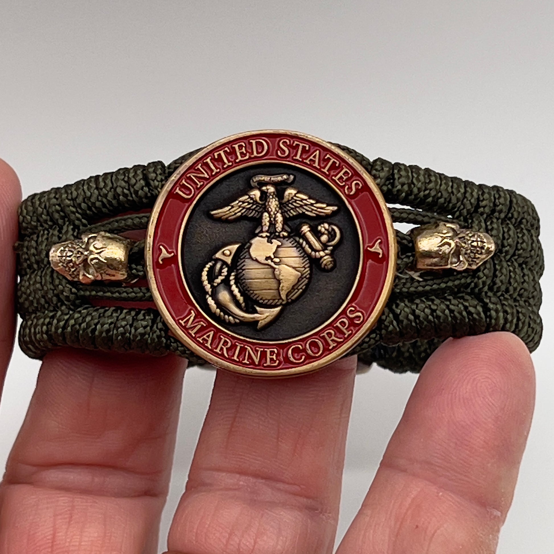 Thin, medium width, flexible and comfortable braided United States Marine Corps bracelet featuring a 24K antique gold plated USMC officer Eagle Globe and Anchor medallion, two gold plated skulls, a gold Cobra style tactical buckle and woven with Olive Drab Type-1 paracord.