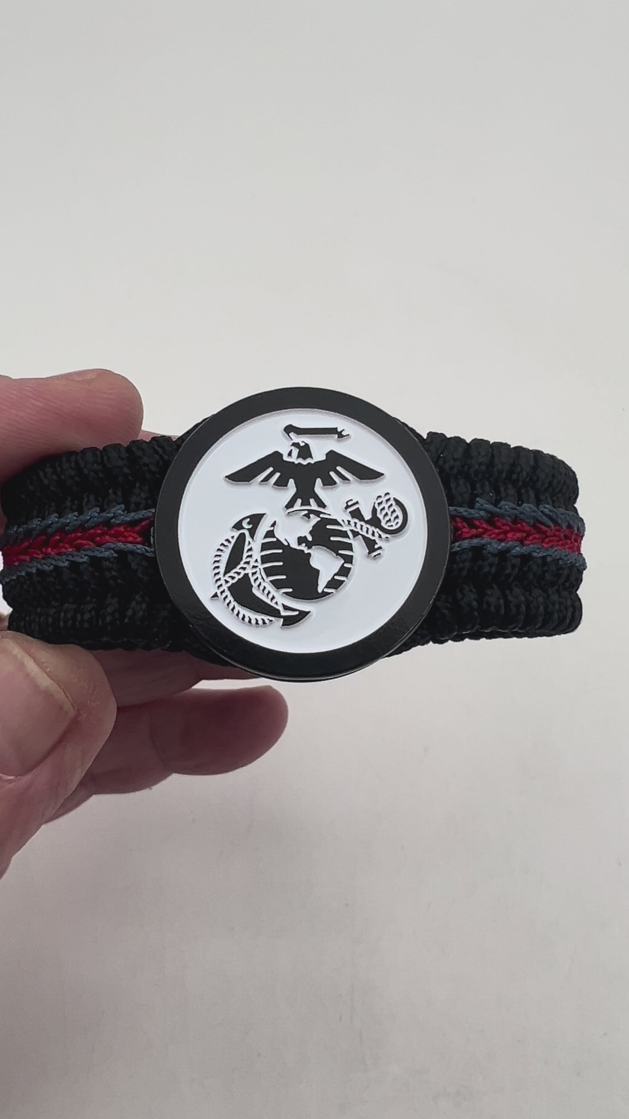 Thin, medium width, flexible and comfortable braided United States Marine Corps bracelet featuring a black USMC Eagle Globe and Anchor medallion, a black Cobra style tactical buckle and woven with black Type-1 paracord and stitched with red and blue nano cord.